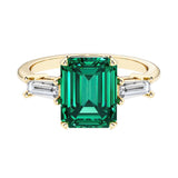 Emerald cut 3 stone engagement ring with green gemstone and diamond accents.