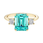 Emerald cut 3 stone engagement ring with green gemstone and diamond accents.