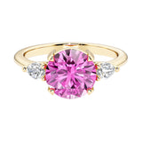 A 14k yellow gold three stone round engagement ring with a brilliant round pink sapphire center diamond and two round side stones.