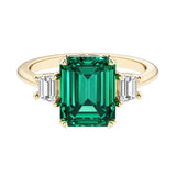 Emerald cut 3 stone engagement ring with green gemstone and diamond accents.