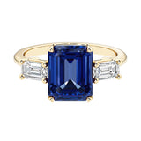 Emerald cut 3 stone engagement ring with blue gemstone and diamond accents.