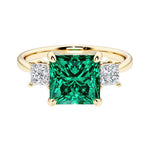 Princess cut 3 stone engagement ring with square side diamonds and green emerald gemstone center.